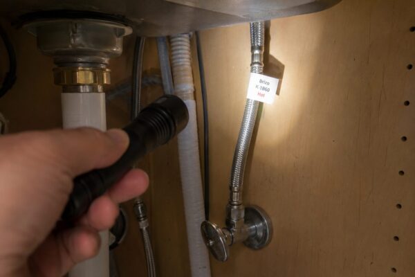 Close-up under-sink view showing braided supply lines connecting to a faucet, where Delta and some Moen faucets keep their model tag