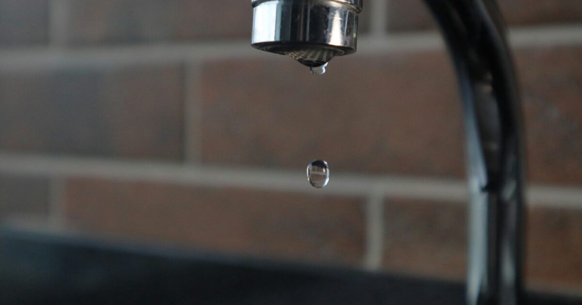 Faucet repair guide hero — kitchen faucet caught mid-drip with a water droplet falling toward the basin
