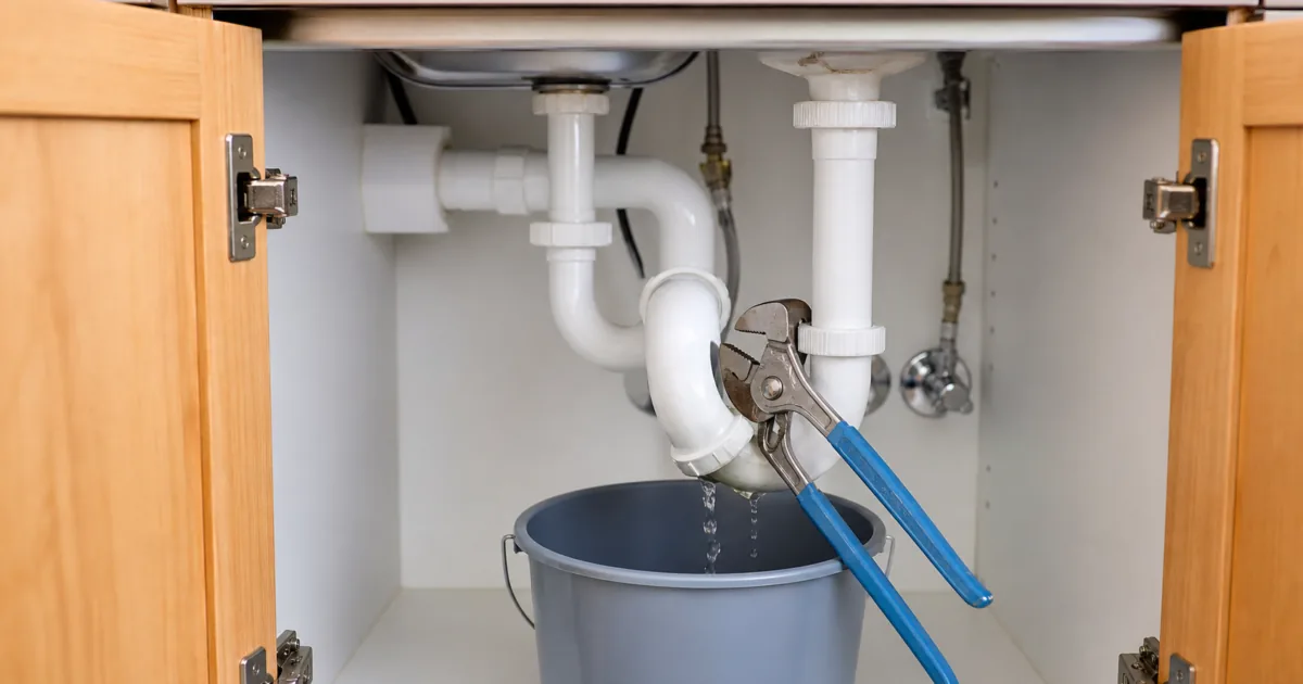 Drain repair guide hero: a homeowner clearing an under-sink P-trap into a bucket with channel-lock pliers